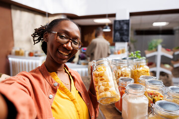 In bio-food shop, black woman wearing spectacles holds glass container loaded with spaghetti. Organic products, vlogging, digital camera, and customer videography are all examples of online marketing.