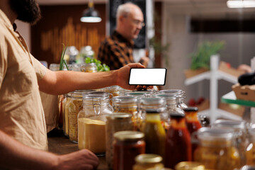 Man in zero waste shop using mockup mobile phone to analyze products. Vegan customer thoroughly checking local supermarket food items are suitable for his diet with isolated screen smartphone - obrazy, fototapety, plakaty