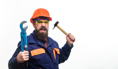 Angry builder in hard hat with adjustable pipe wrench and hammer. Bearded man in construction uniform and protective helmet with adjustable spanner and hammer. Plumber with monkey wrench and hammer.