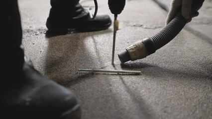 One man drills a hole in the floor with a hammer drill, the second holds a vacuum cleaner nearby. Floor repair, drilling the floor to install dowels.