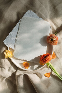 Overhead view of handmade deckle edge paper on a ceramic plate with orange ranunculus flowers on a linen tablecloth in sunlight