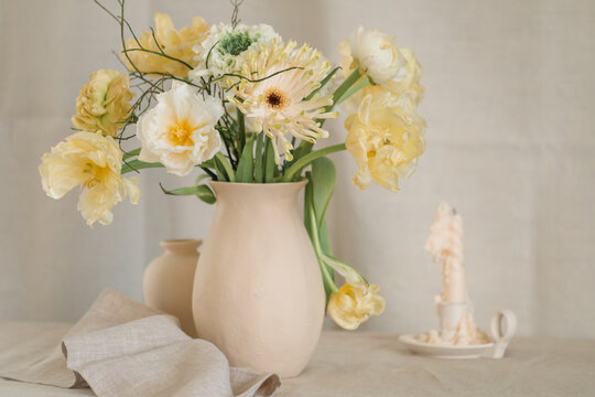Bunch of yellow gerbera, tulip and ranunculus flowers in a vase on a table with a candle and napkin