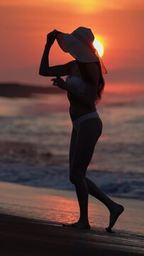 Woman in white bikini and straw sun hat walking on sandy beach at sunrise. Barefoot female enjoyment during summer beach holiday at golden hour on morning. Slow motion, handheld shot, vertical video