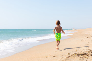 Fototapeta premium Cute little boy playing on the beach on summer holidays. Joyful child in nature with beautiful sea, sand and blue sky. Happy kid on vacations at seaside running in the water and having fun