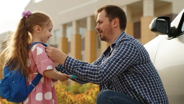 Smiling father giving textbook daughter pupil with backpack hugging before going school campus closeup. Happy dad embracing little female kid child arriving elementary education lesson at schoolyard