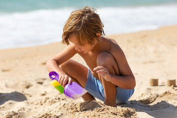 Happy child little boy playing with the sand on the beach, building sand castle, enjoying summer, playing. Advertising for tour operators, hotels, airlines. Vacation concept. - obrazy, fototapety, plakaty
