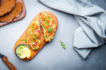 Avocado sandwich with salmon on rye bread with guacamole sauce, arugula and sesame seeds on wooden board on gray table background, top view