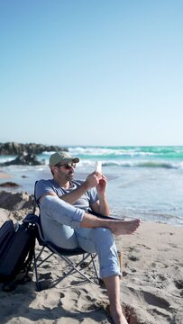 mature man digital nomad sitting alone on the beach relaxing taking photos with his cell phone