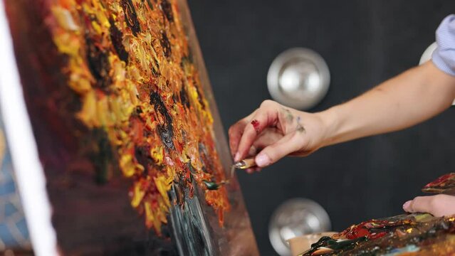 Close up view female artist hands with palette knife and color paints on the palette. Painter at the studio creating still life with sunflower on canvas using oil paintings and palette knife.