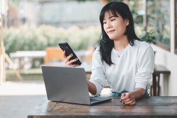 Contented Asian woman multitasking with a smartphone and laptop at a rustic wooden table in a well-lit cafe space.