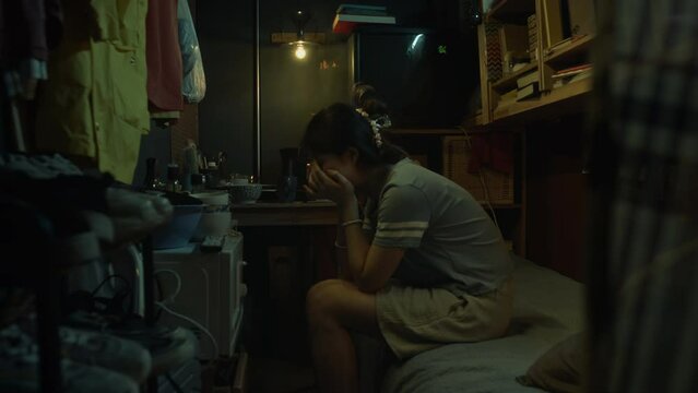 Medium full footage of sad young Chinese woman sitting on bunk bed in cramped dark shoebox apartment, clutching face in hands and crying in despair at hopeless poverty