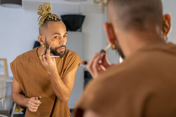 Black male gay applying make up looking mirror standing back to camera. Stylish homosexual man in...
