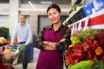 Asian woman greengrocer worker in uniform standing in salesroom and looking in camera. Man with...