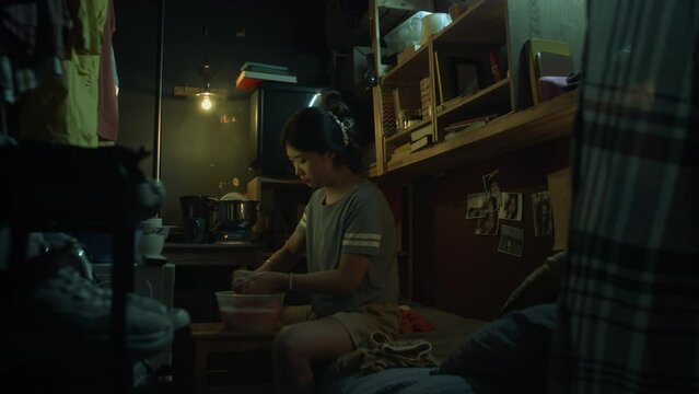 Full shot of young Chinese woman in t-shirt and shorts sitting on bed in dimly lit cluttered micro apartment at nighttime, washing clothes by hand in plastic tub of soapy water