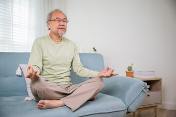 Lifestyle senior man sitting on sofa in living room holding hands in mudra practicing home yoga in...