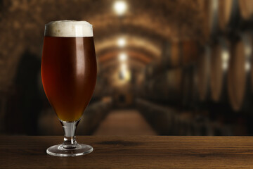 Glass with fresh beer on wooden table in cellar, space for text