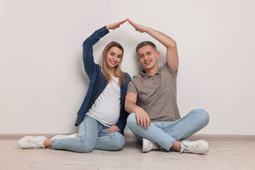 Young family housing concept. Pregnant woman with her husband forming roof with their hands while sitting on floor indoors