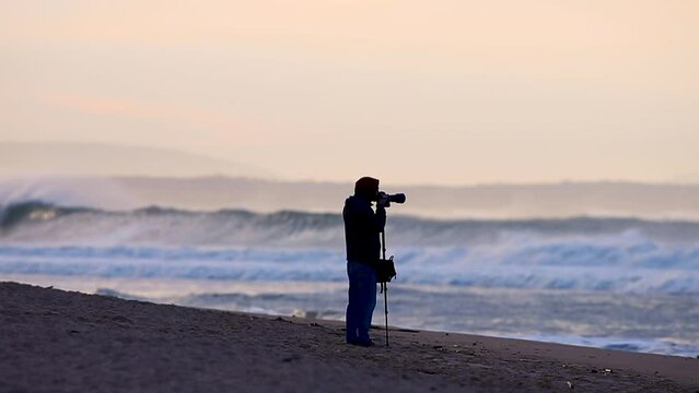 Photographer with a tripod takes pictures of a Portuguese beach at sunrise. Dark silhouette of a man and waves crashing on the beach