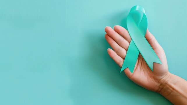 A hand gently holds a teal ribbon against a soft turquoise background symbolizing ovarian cancer awareness