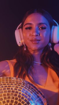 Vertical shot of female Asian zoomer in headphones partying with disco ball in neon nightclub