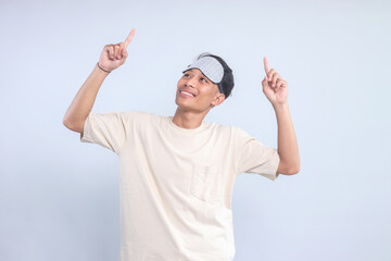 Happy Asian man wear sleeping mask, looking and pointing up to the copy space with both hands 