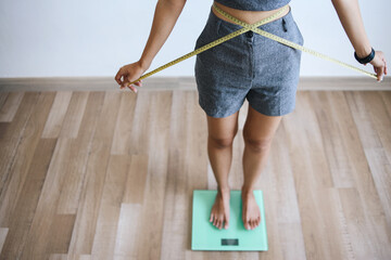 Slim woman standing on weigh scales while measuring her waist by measure tape.
