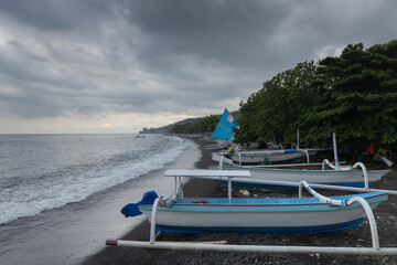Black sand beach, fihsermen, boats and overcast sky at coastline of touristic Amed village in Bali...
