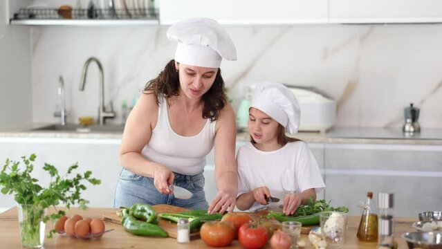 Mom and daughter in hats dice up raw vegetables for salad on cutting boards. Girl listens attentively to her mother, carefully works with knife, tries to follow recommendations