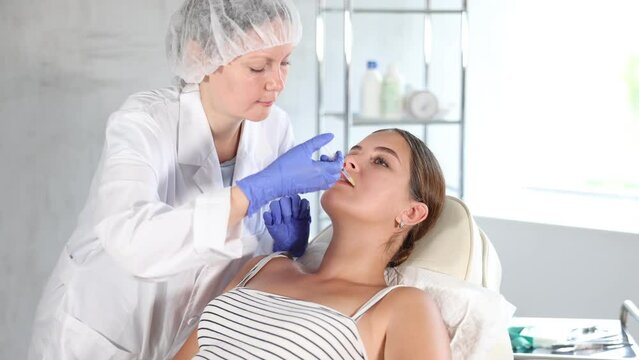 Unrecognizable circumcised female cropped doctor in disposable medical mask manipulates introduction of filler and correction of shape and size of lips to girl patient. Close-up