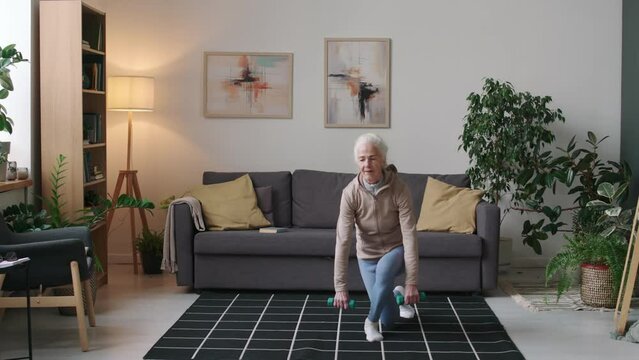 Full stab shot of fit Caucasian retired woman in activewear doing diagonal lunge exercise with dumbbells at home