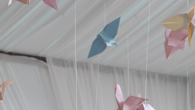 Close, slow motion footage of colourful pastel origami paper crane birds floating in breeze, hung from roof, blue, yellow, pink and orange.
