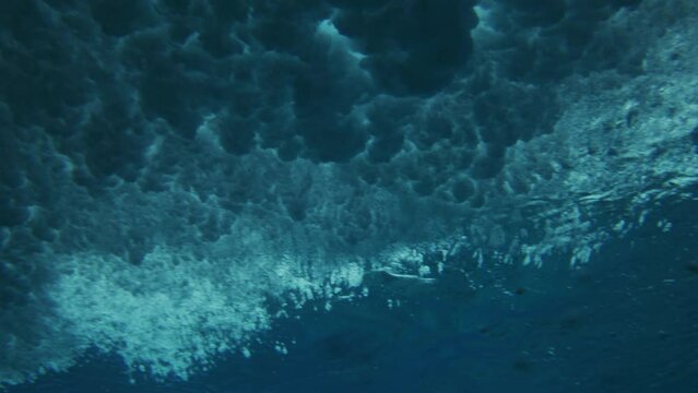 Breaking wave crashes as bubbles spread across ocean surface twirling in vortex, underwater angle