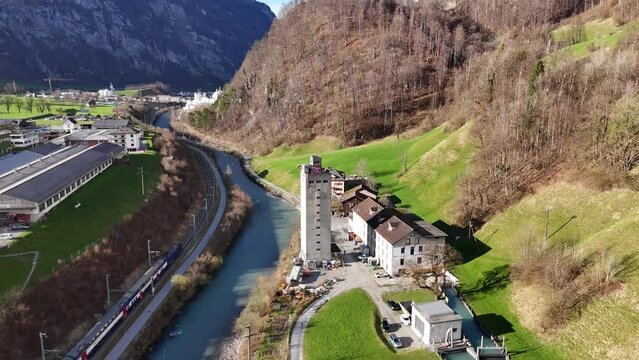 Aerial approaching shot of tower and rolling mill factory beside river and riding train. Glarus City, Switzerland. Drone descending shot.