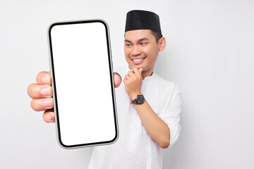 Smiling young Asian Muslim man showing smartphone screen, touching his chin, thinking about the offer on white background. People religious Islam lifestyle concept. celebration Ramadan and ied Mubarak