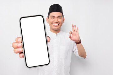 Smiling young Asian Muslim man showing display mobile phone screen, showing okay gesture isolated on white background. People religious Islam lifestyle concept. celebration Ramadan and ied Mubarak