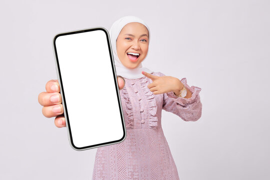 Cheerful beautiful young Asian Muslim woman wearing hijab and purple dress pointing index finger at empty smartphone screen isolated on white studio background