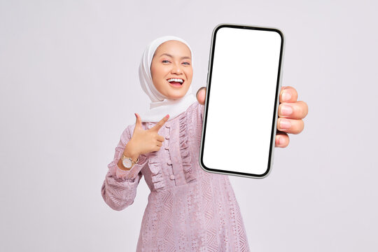 Cheerful beautiful young Asian Muslim woman wearing hijab and purple dress pointing index finger at empty smartphone screen isolated on white studio background