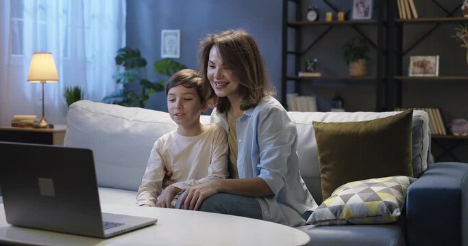 Smiling woman and kid talking to webcam, video conference call. Mother and son waving hands, sharing news during online video chat sitting on the couch in the evening