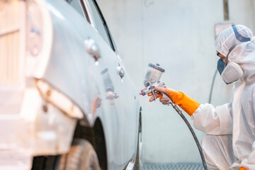 Car painter in protective clothes and mask painting automobile bumper with metallic paint and varnish in chamber workshop