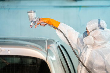 Car painter in protective clothes and mask painting automobile bumper with metallic paint and...