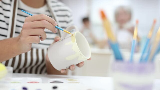 Closeup of hands of talented female artisan creating own unique ceramic items in pottery workshop, painting mug with engobe and brush. Craft activity concept