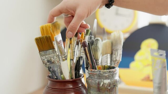 Artist picks up paint brush. Female artist hand picking up brush for painting. Unrecognizable young artist choosing brushes. Tools for painting. Close-up in 4K, UHD