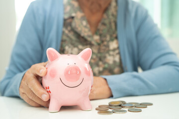 Asian elderly woman putting coin into pink piggy bank for saving money and insurance, poverty, financial problem in retirement.