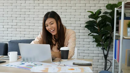 Foto op Canvas Asian Female freelance using laptop at home office desk. Woman reading financial graph chart Planning analyzing marketing data. Asian female people working office firm with business stuff, coffee cup © aFotostock