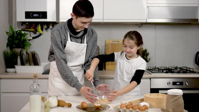 Happy and smiling girl helps brother in kitchen. Teenage children whisking egg in bowl with metal wire whisk. Kids pastry chef preparing dough. Cooking process cookies and pastry
