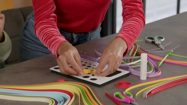 Close-up of black female hands rolling colorful paper strip and attaching with pin on board, while making decorative designs and teaching child to quilling indoors.