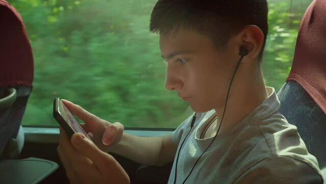 Close up. Boy teenage using smartphone while sitting near window in public transport shuttle bus playing online game. Writes message listens to music through wired headphones in train. Excursion