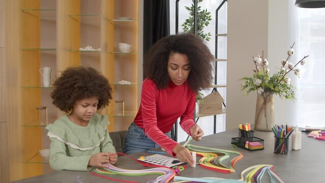 Positive lovely school age curly African American daughter and beautiful black mother making paper quilling designs together, developing artistic and creative kid skills while relaxing at home.