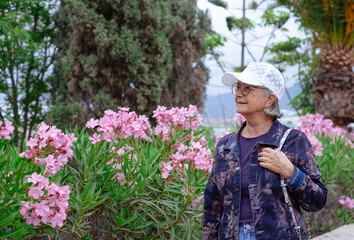 Serene senior woman in denim jacket and white cap walking outdoors in the park,  pink oleander...