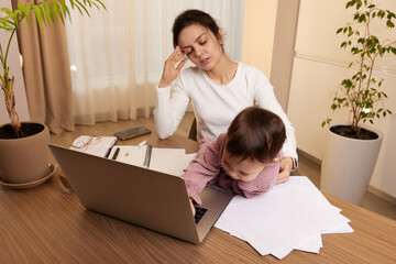 Stressed woman working on laptop at home with her little baby girl. Child makes noise and disturbs mother at work.